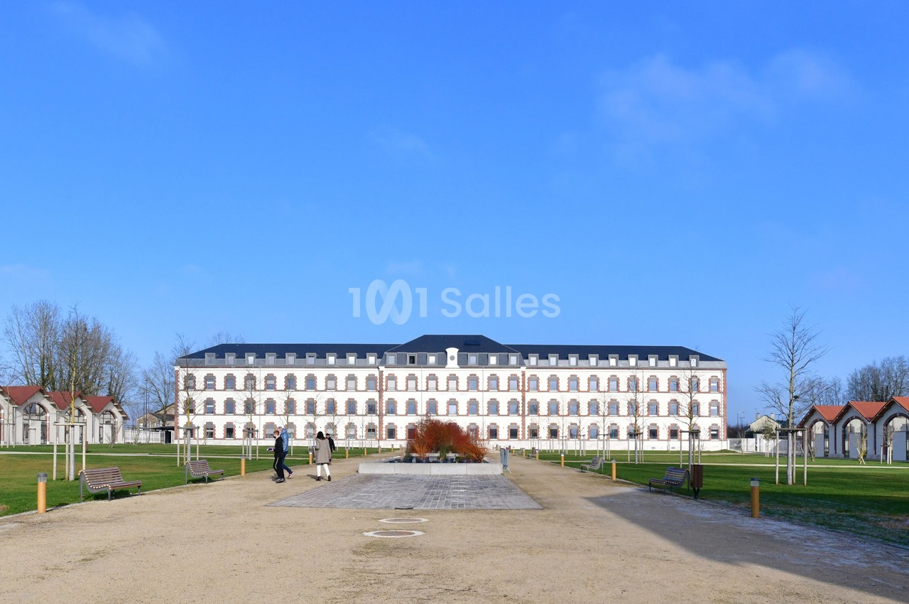 Bâtiment historique en pierre avec des fenêtres régulières, entouré d'espaces verts et de bancs sous un ciel bleu.