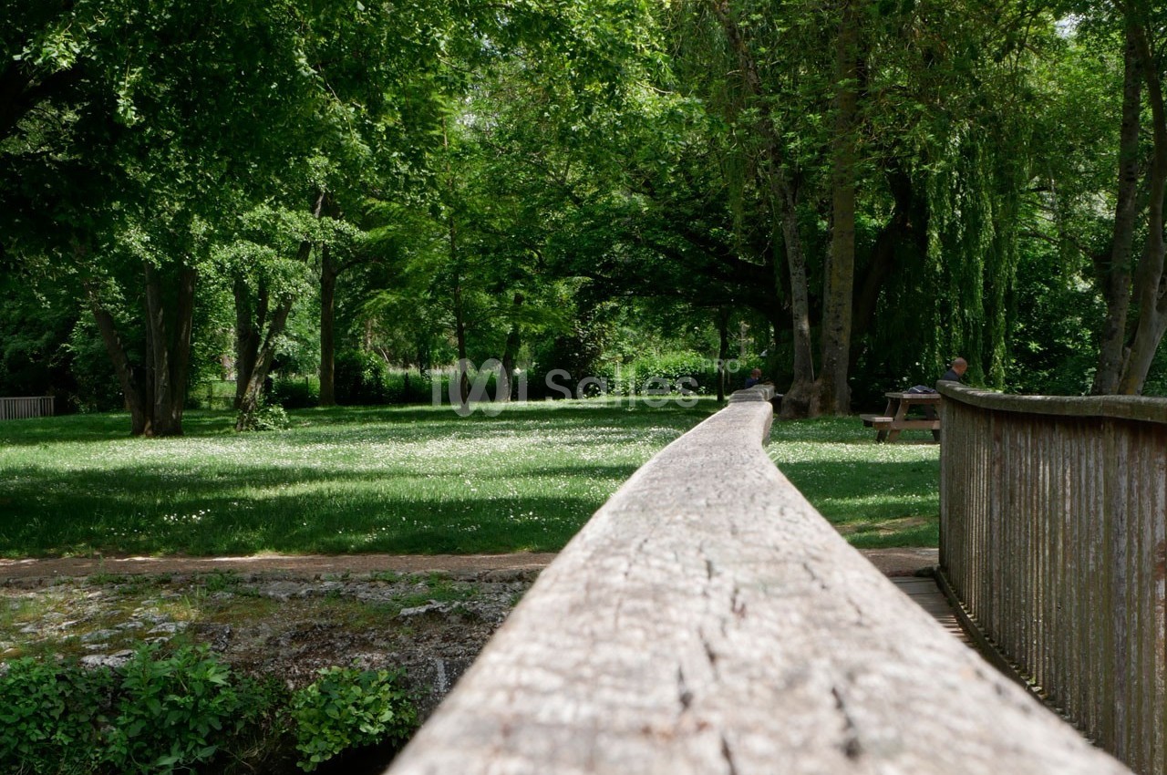 Vue d'un parc verdoyant avec un pont en bois au premier plan et des arbres ombrageant une pelouse.