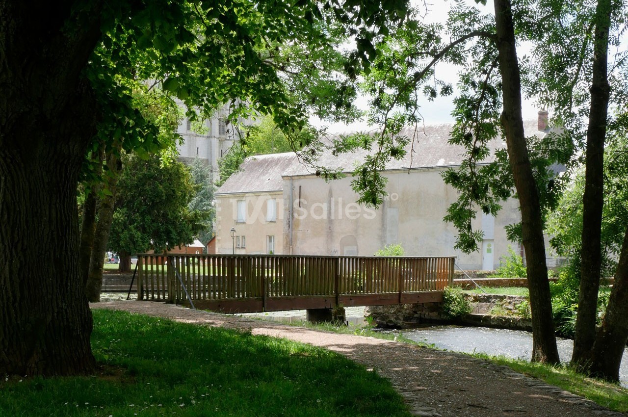 Petit pont en bois traversant un cours d'eau, entouré d'arbres et donnant sur des bâtiments anciens en arrière-plan.
