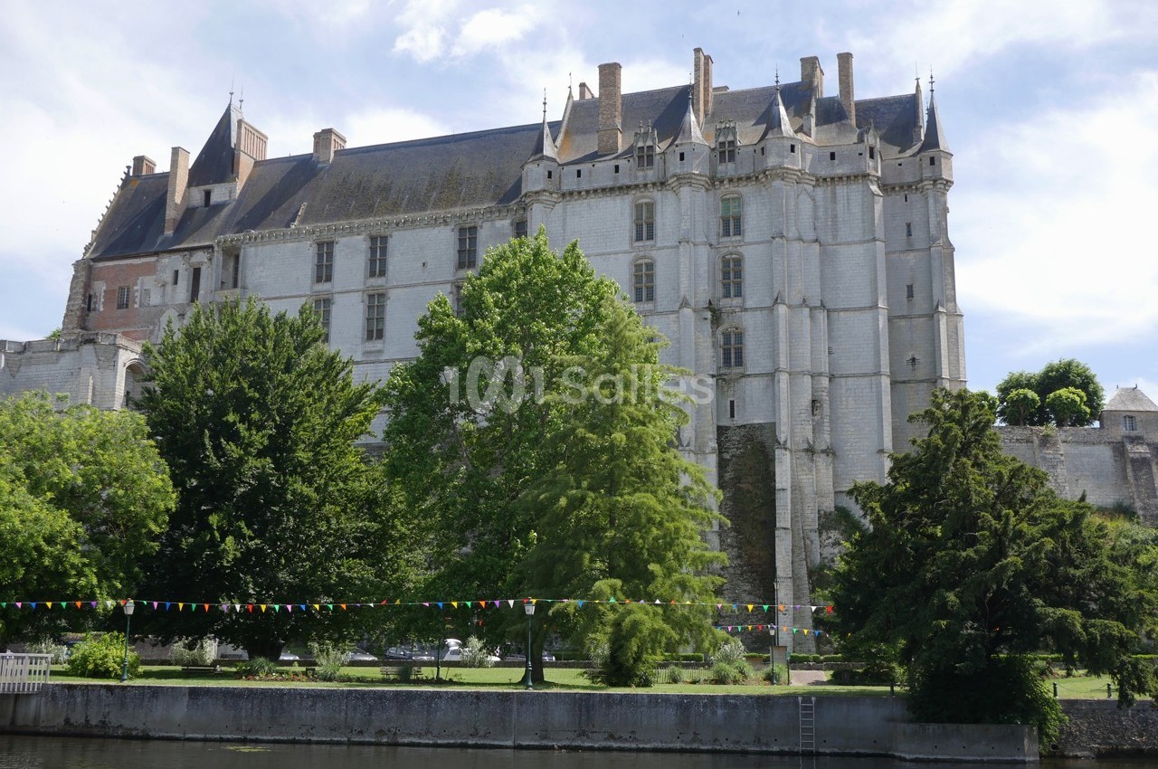 Façade d'un château médiéval en pierre, entouré d'arbres et bordé d'une rivière avec des guirlandes colorées.