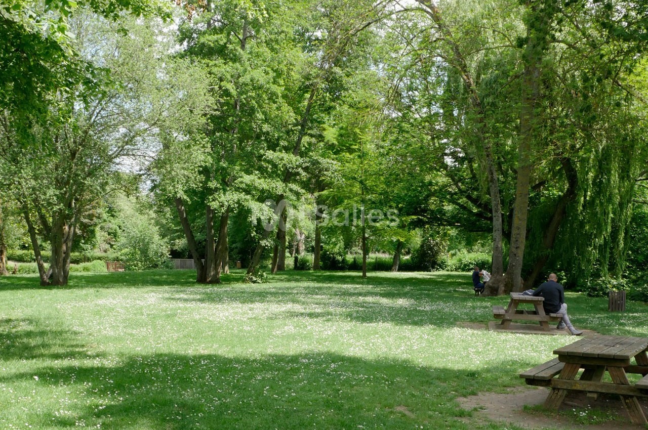 Espace vert avec pelouse, arbres et tables de pique-nique, deux personnes assises dans un cadre calme et ombragé.