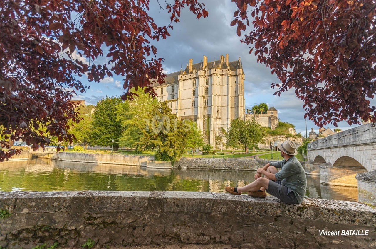 Un homme assis sur un muret admire un château médiéval entouré d'arbres, près d'une rivière et d'un pont en pierre.