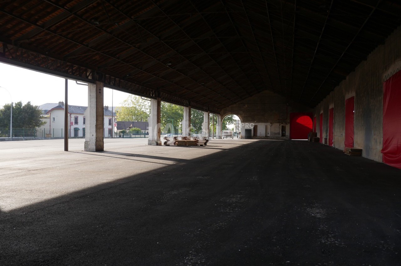 Hangar ouvert avec une structure métallique, sol asphalté, lumière naturelle et bâtiments visibles à l'extérieur.