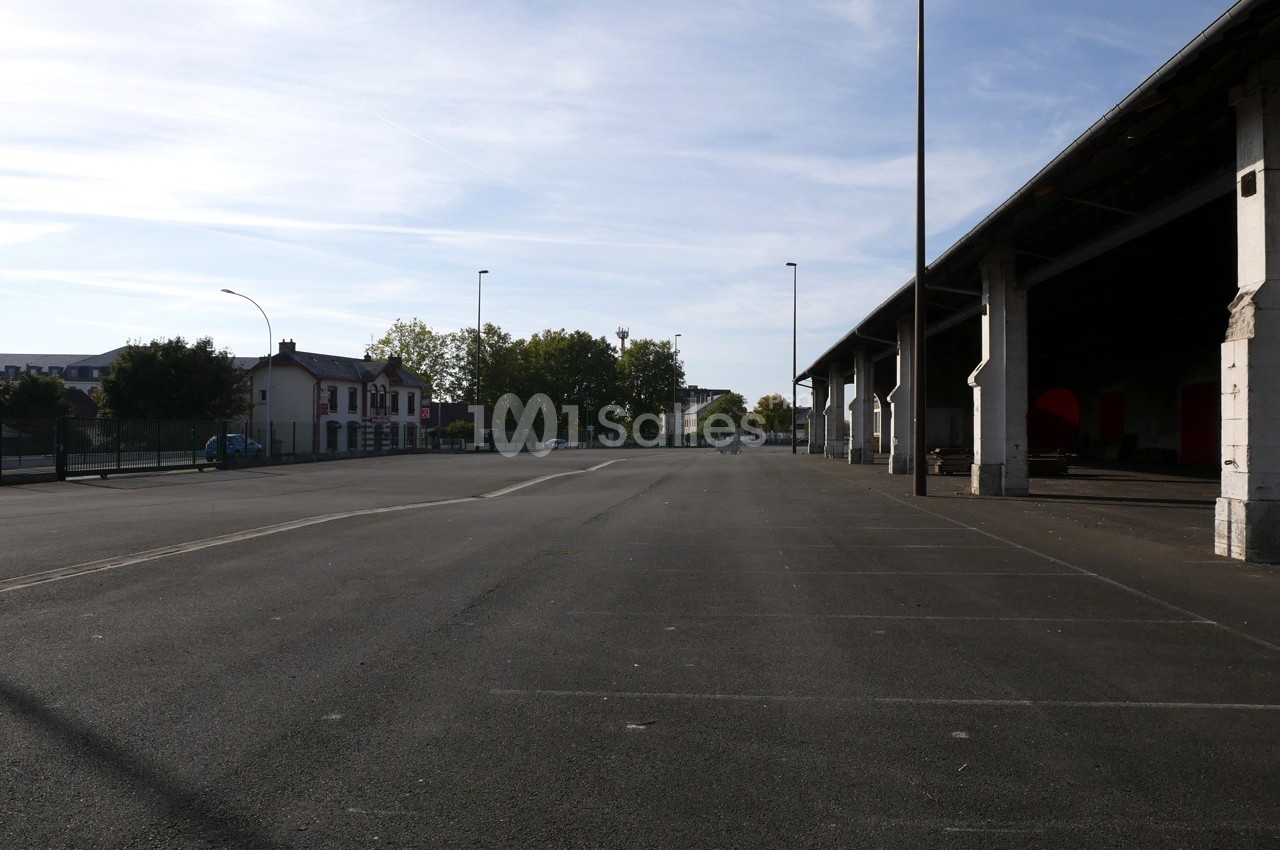 Grand parking vide avec un bâtiment couvert sur la droite et quelques arbres en arrière-plan sous un ciel dégagé.