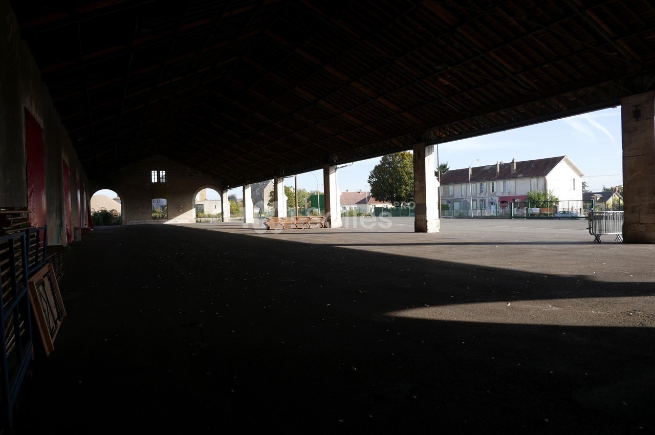 Vue d'un grand hangar ouvert avec une cour pavée et des bâtiments résidentiels en arrière-plan.
