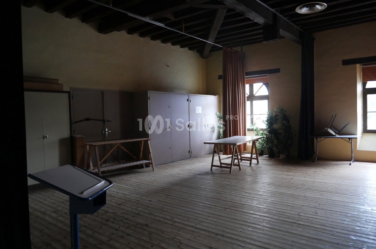 Salle vide avec parquet en bois, meubles de rangement, table, chaises empilées et lumière naturelle provenant des fenêtres.