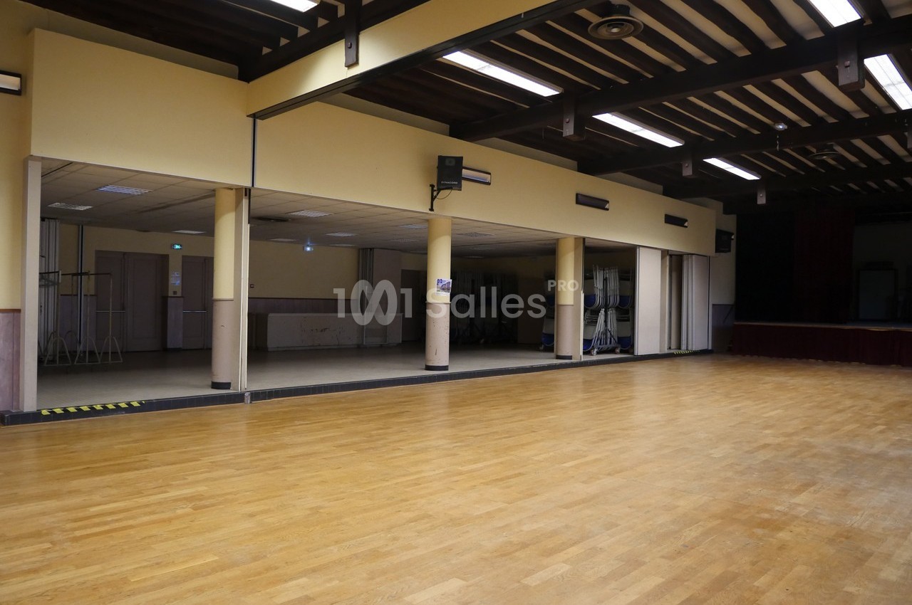 Salle spacieuse avec parquet en bois, poutres apparentes au plafond et scène en arrière-plan.