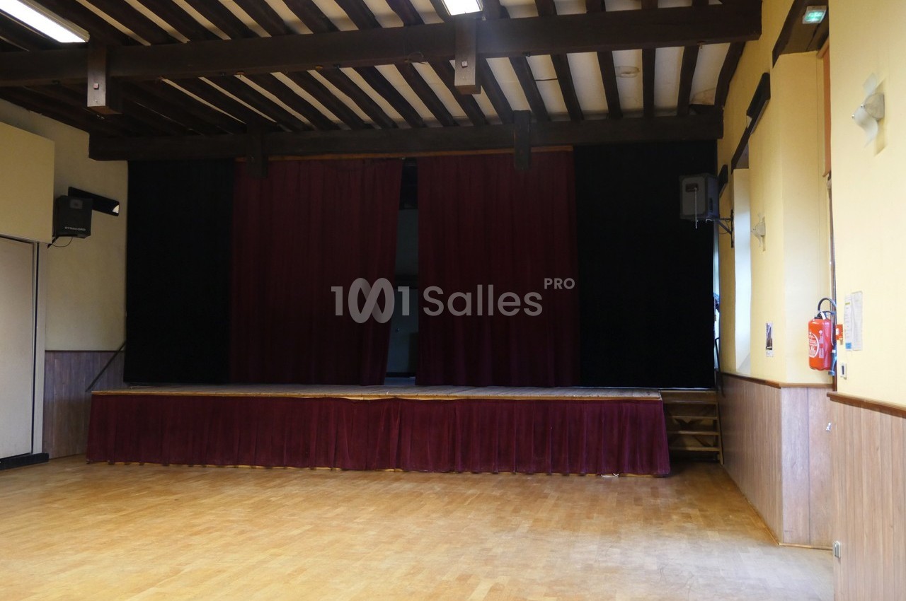 Scène de petite salle avec rideaux rouges fermés, parquet au sol et poutres apparentes au plafond.