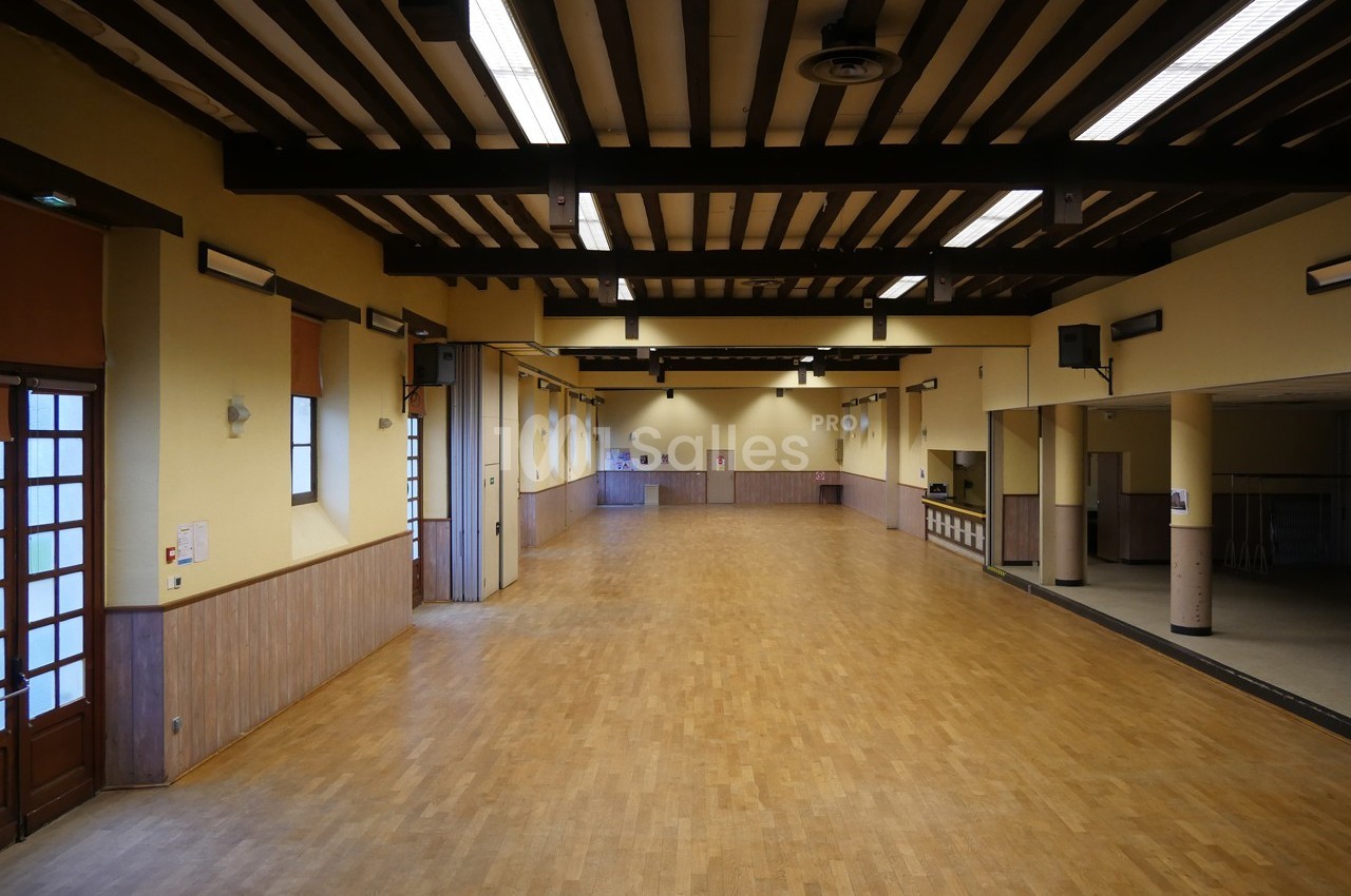 Salle spacieuse avec parquet en bois, murs jaunes, poutres apparentes et éclairage fluorescent.