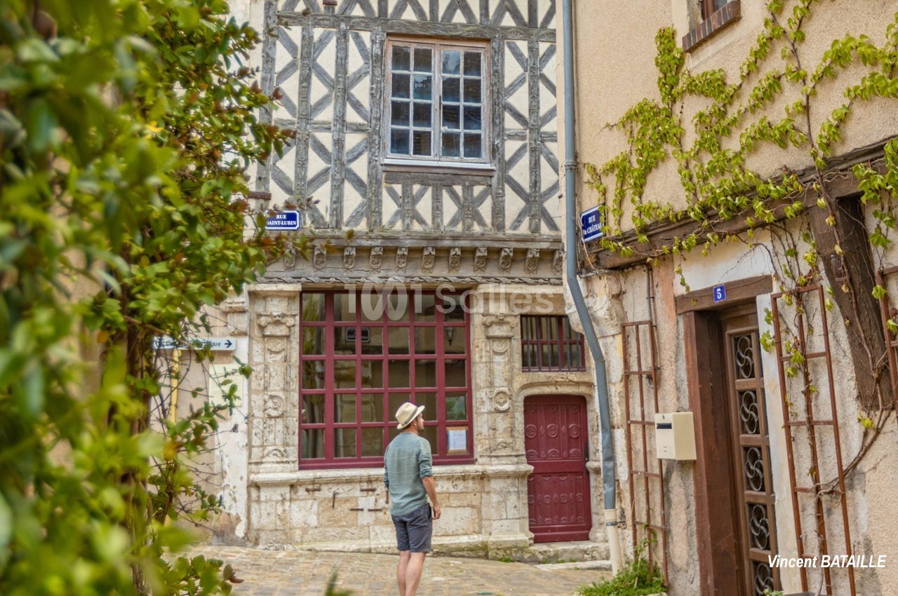 Homme de dos marchant dans une ruelle pavée bordée de façades anciennes, dont une maison à colombages.