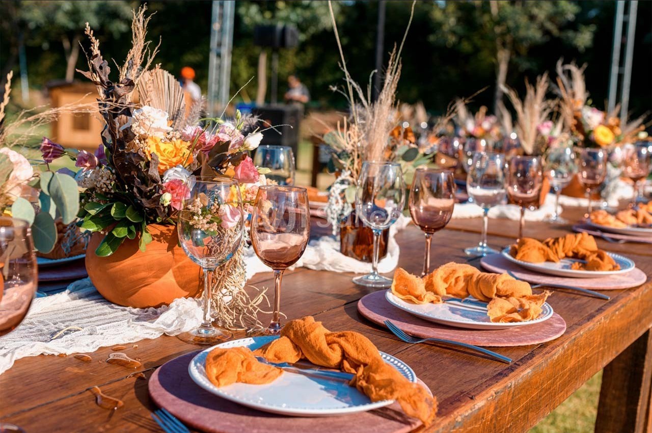 Table en bois décorée pour un repas en extérieur, avec fleurs séchées, verres colorés et serviettes orange.