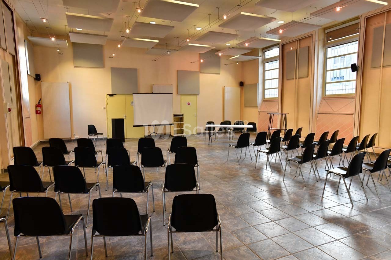 Salle de réunion lumineuse avec des rangées de chaises noires, un écran de projection et des tables à l'avant.