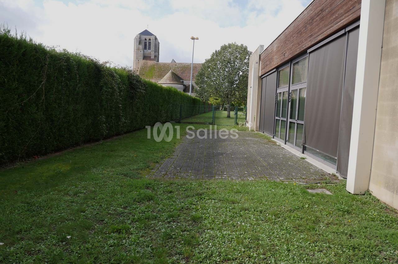 Allée enherbée bordée de haies et d'un bâtiment moderne, avec une église en arrière-plan sous un ciel dégagé.