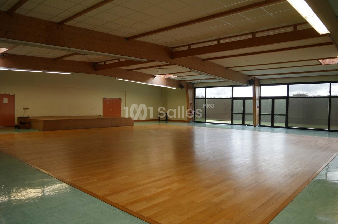 Salle polyvalente vide avec parquet en bois, murs clairs, grandes baies vitrées et plafond avec poutres apparentes.