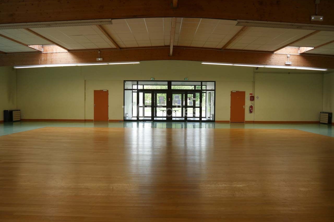 Salle vide avec parquet au sol, murs clairs, plafond en poutres apparentes et portes vitrées donnant sur l'extérieur.