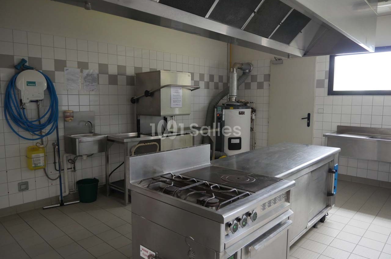 Cuisine professionnelle avec plan de travail en inox, cuisinière, évier, hotte et équipements muraux dans un espace carrelé.