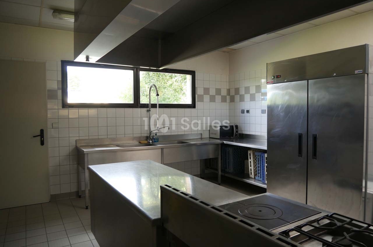 Cuisine professionnelle équipée avec plan de travail en inox, réfrigérateur, évier, plaques de cuisson et fenêtres.