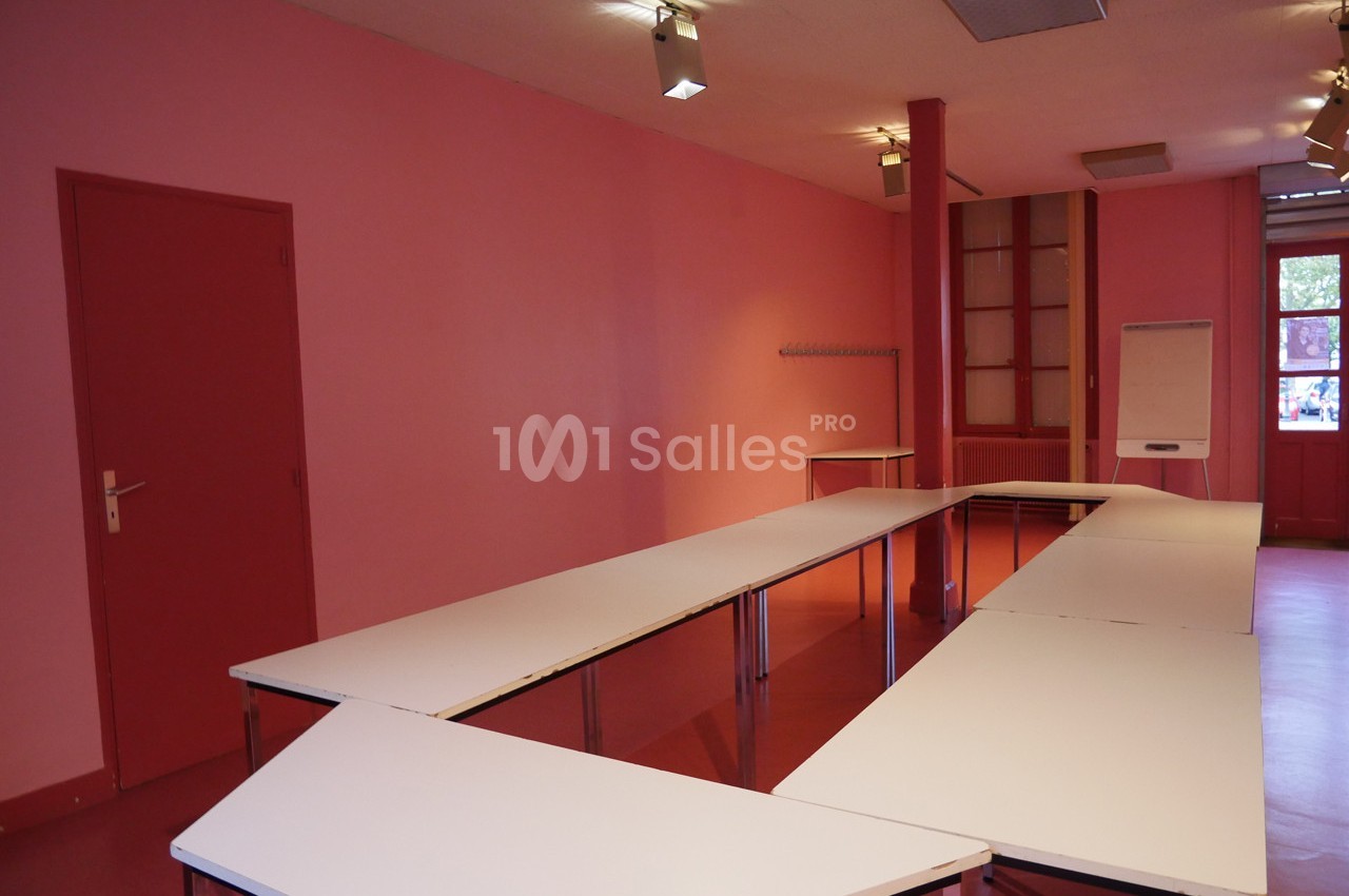 Salle vide avec murs roses, tables blanches disposées en U, tableau blanc et porte rouge en arrière-plan.