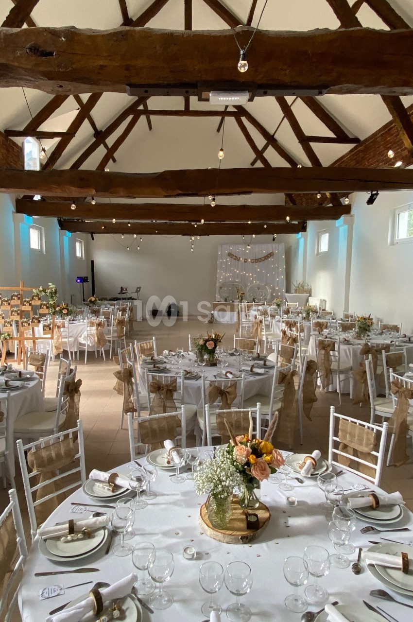 Salle de réception décorée pour un mariage, avec tables dressées, chaises blanches et poutres apparentes au plafond.
