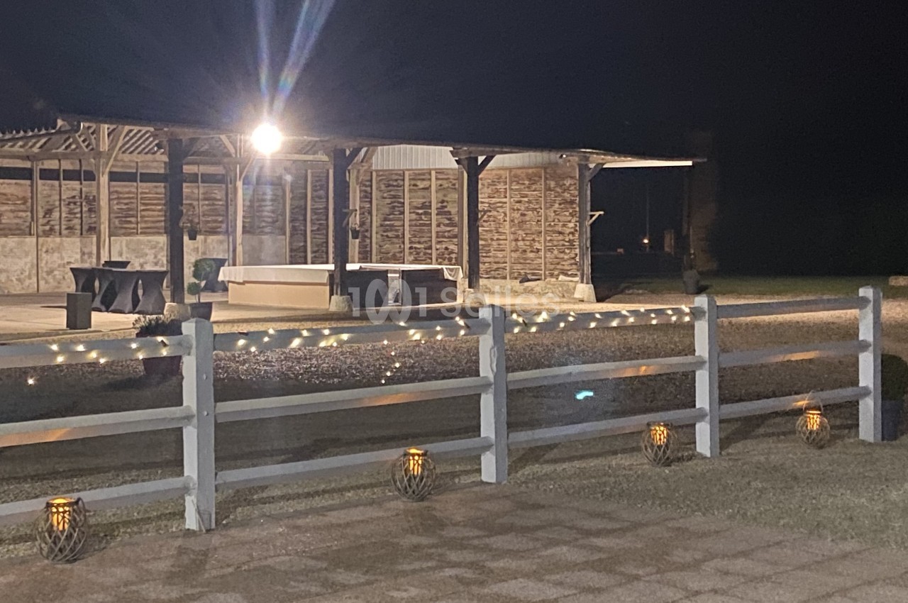 Espace extérieur éclairé de nuit avec une pergola, des guirlandes lumineuses et des lanternes au sol.