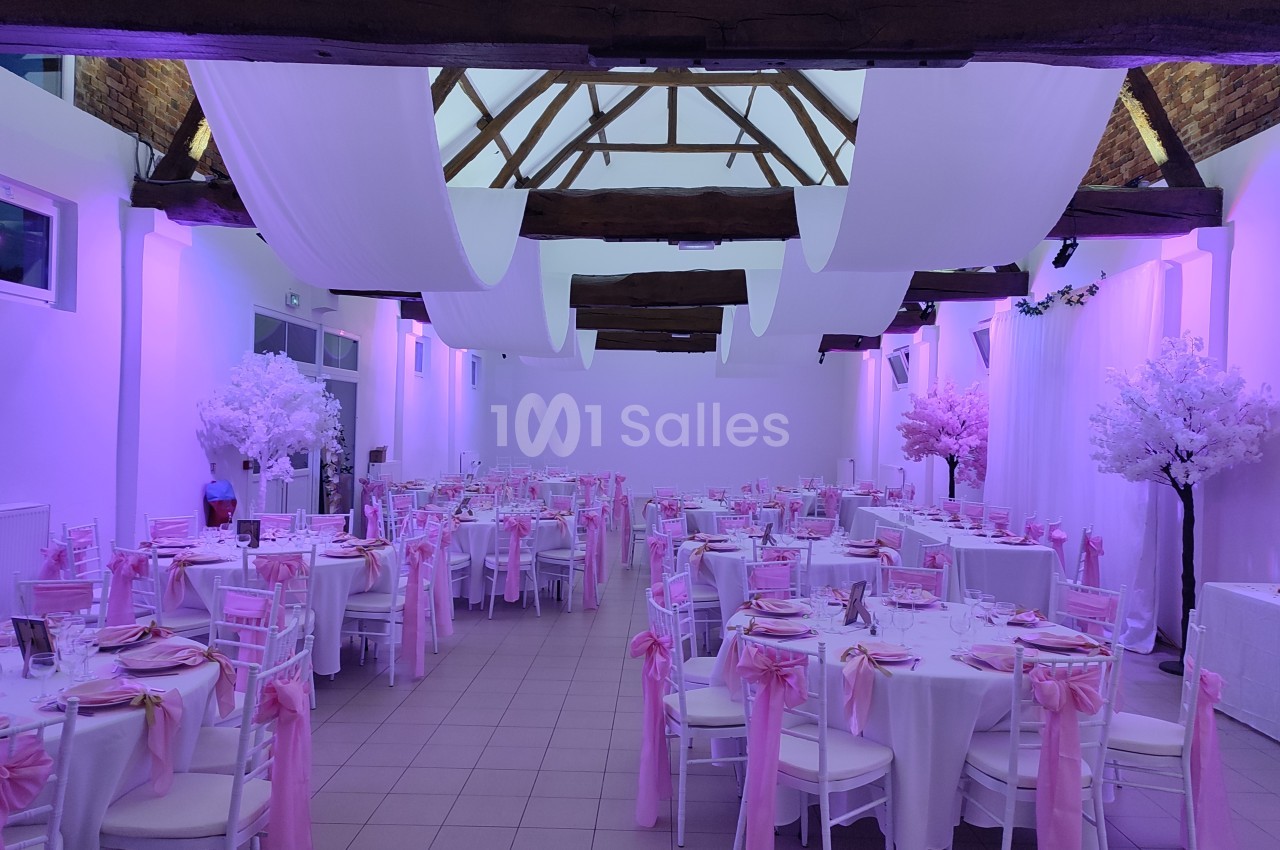 Salle de réception décorée avec des nappes blanches, des chaises ornées de rubans roses et un éclairage violet.