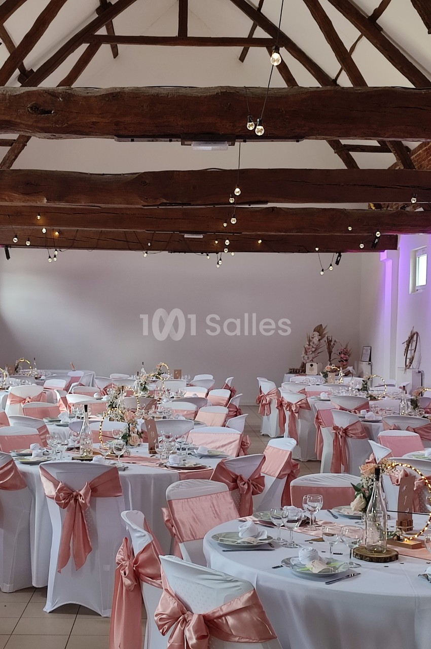Salle de réception décorée pour un mariage, avec des tables dressées et des chaises ornées de nœuds roses.