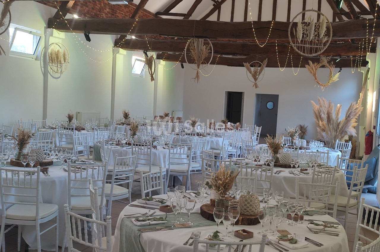 Salle de réception décorée avec des tables rondes, nappes blanches, chaises blanches et ornements végétaux suspendus.