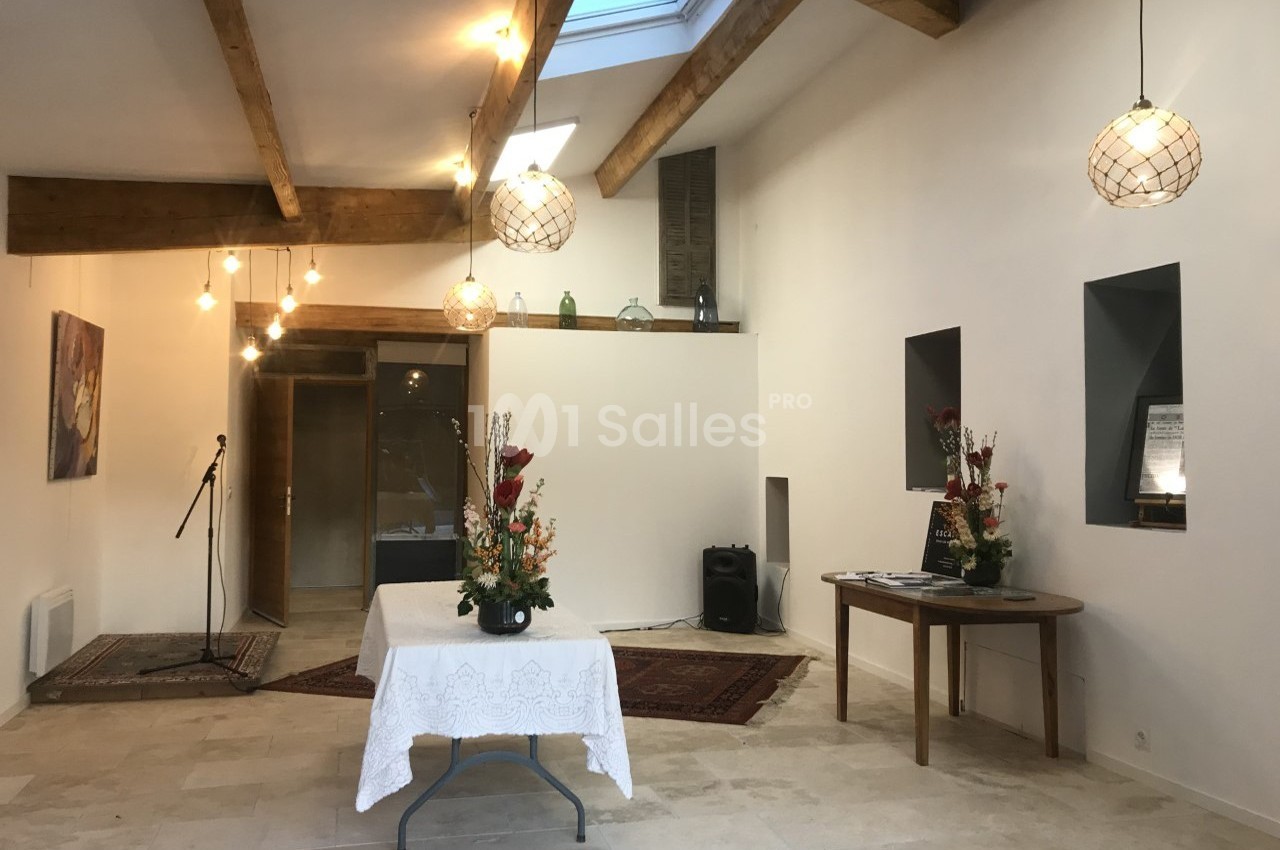 Salle lumineuse avec poutres apparentes, décorée de fleurs sur des tables, éclairée par des suspensions et une verrière.
