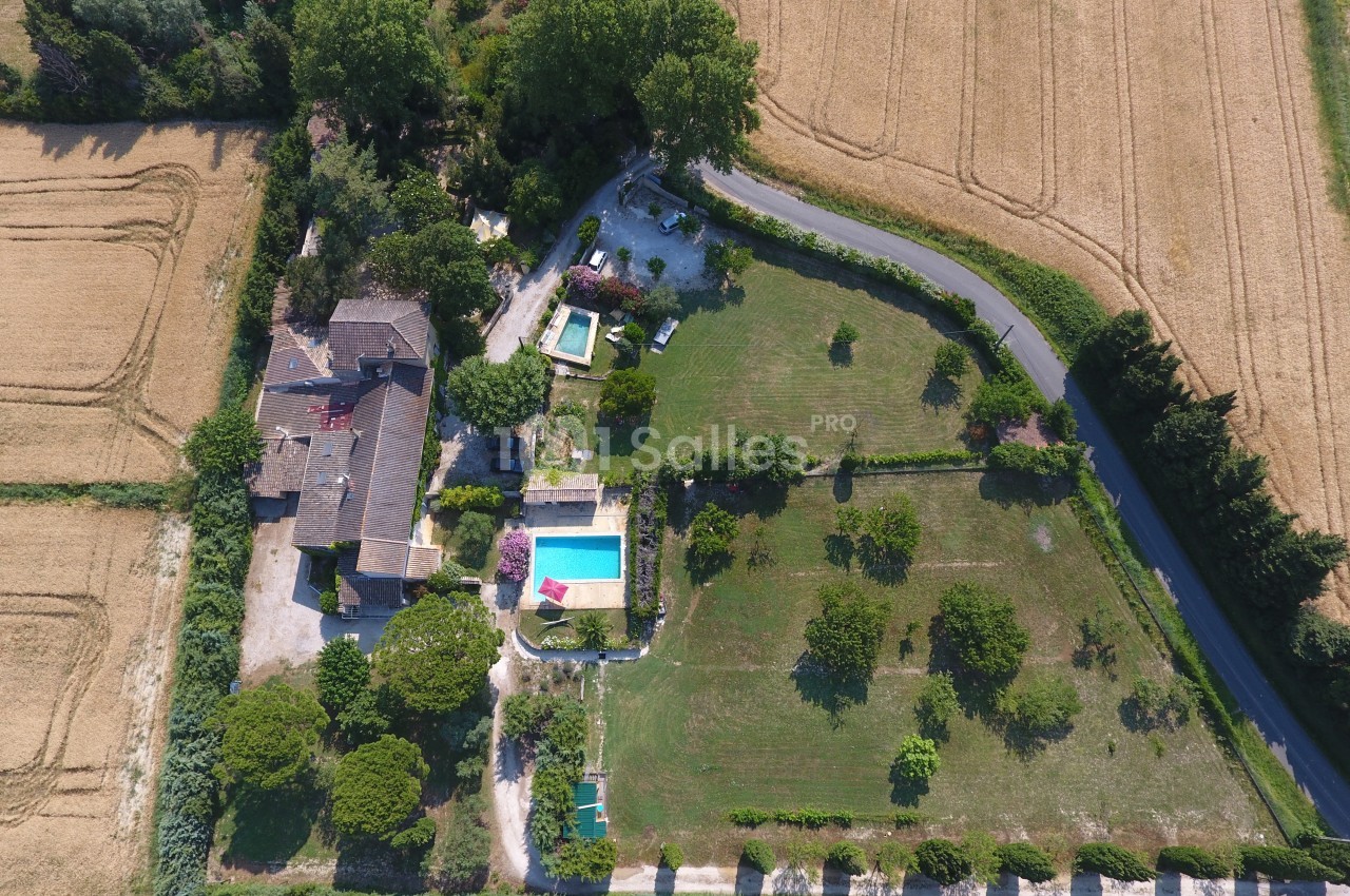 Vue aérienne d'une propriété avec piscine, jardin arboré et champs environnants, bordée par une route de campagne.
