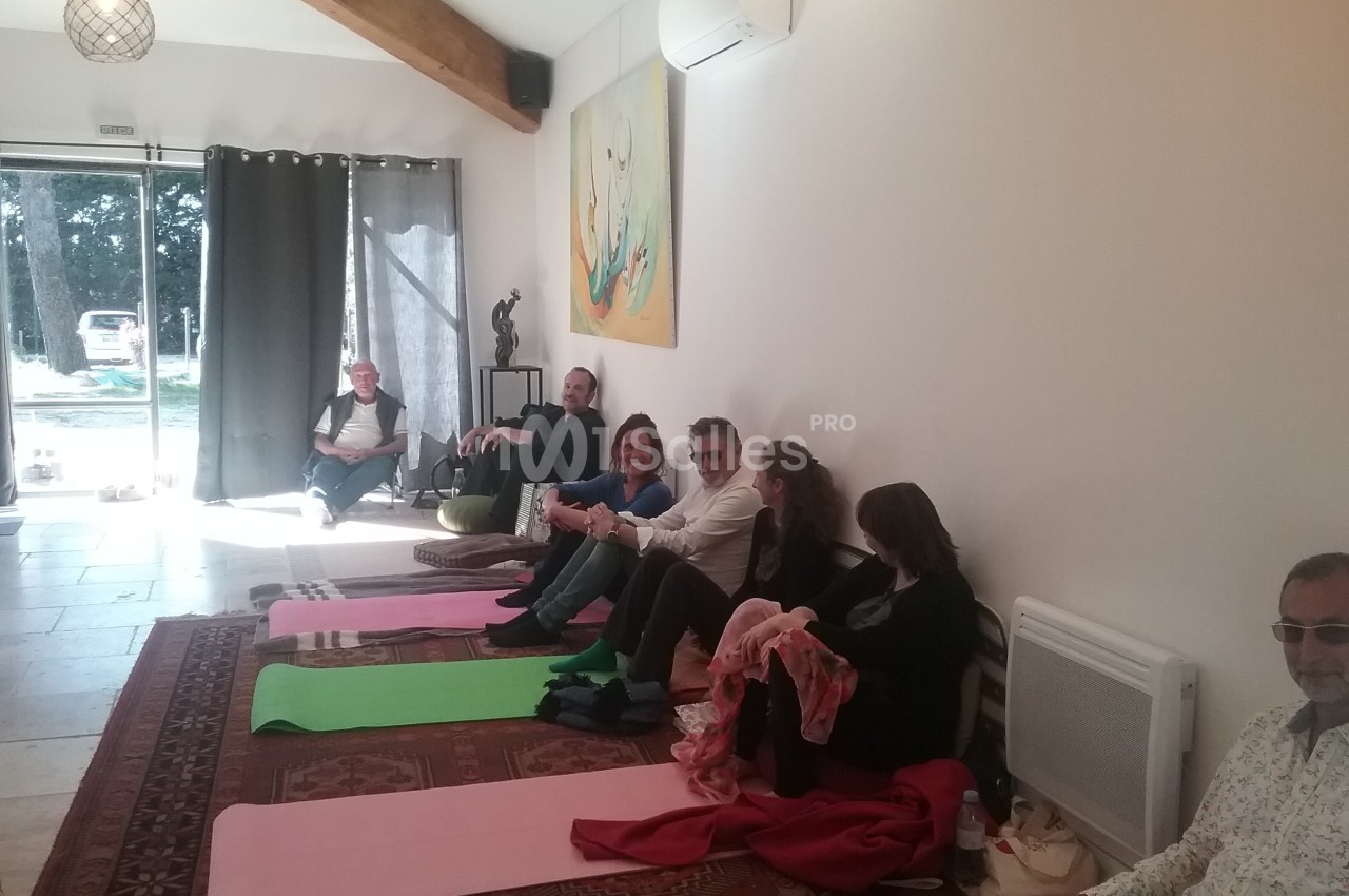 Des personnes assises sur des tapis et des chaises dans une salle lumineuse avec un tableau et des rideaux.