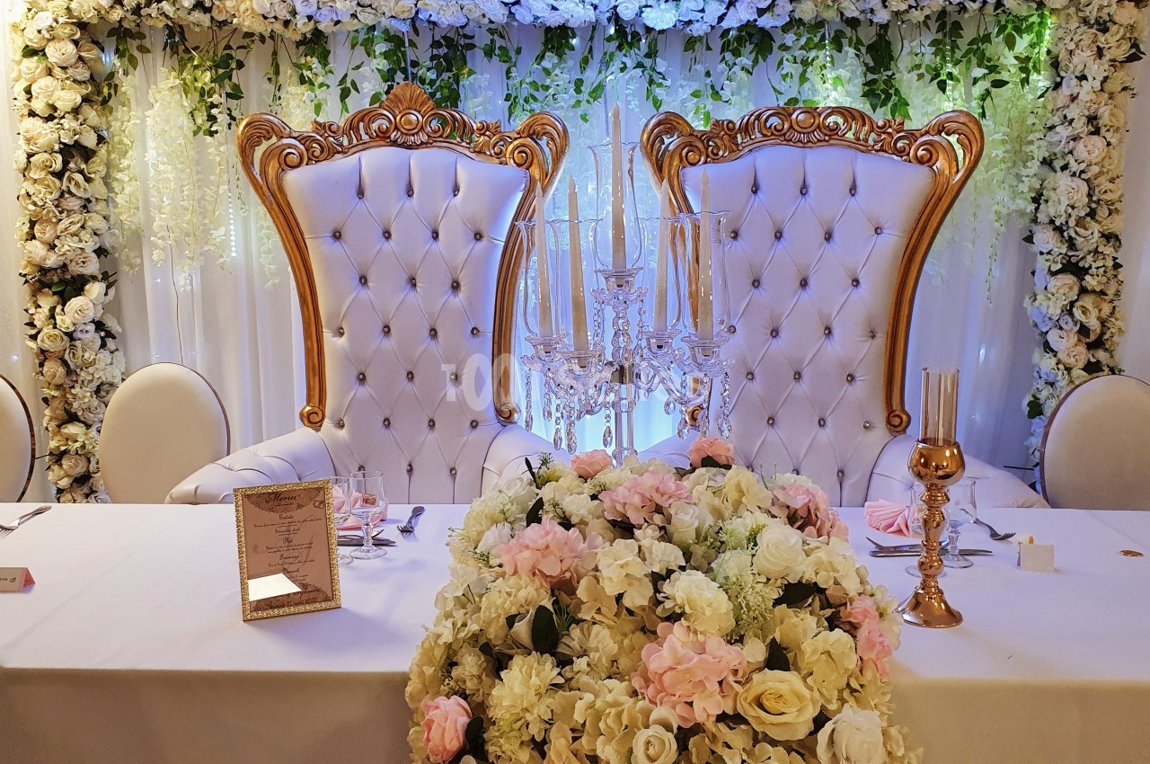 Deux fauteuils ornés dorés devant un mur de fleurs blanches, avec une table décorée de bougies et de fleurs.