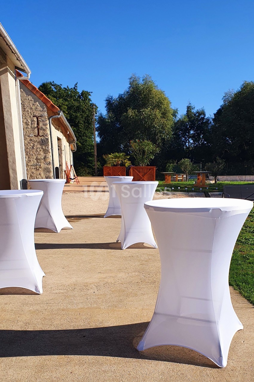 Tables hautes recouvertes de housses blanches disposées sur une terrasse ensoleillée près d'un bâtiment en pierre.