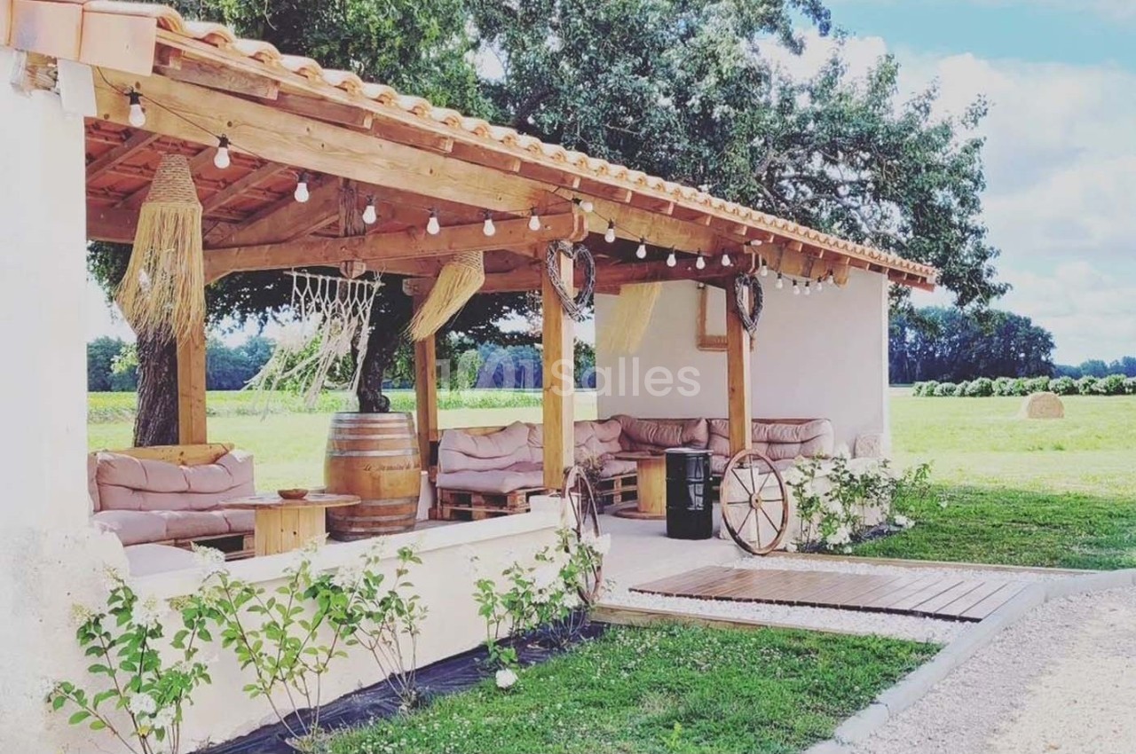 Terrasse couverte en bois avec canapés, décor champêtre, tonneau en table et vue sur un champ verdoyant.