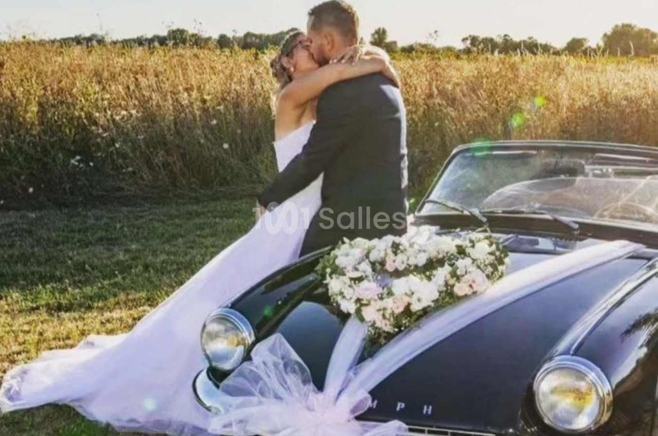 Un couple en tenue de mariage s'embrasse devant une voiture classique décorée de fleurs, dans un champ ensoleillé.