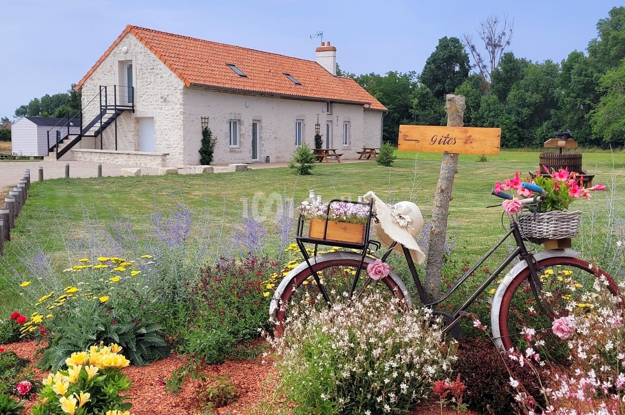 Jardin fleuri avec un vélo décoratif devant une maison en pierre à toit rouge, entourée de verdure.