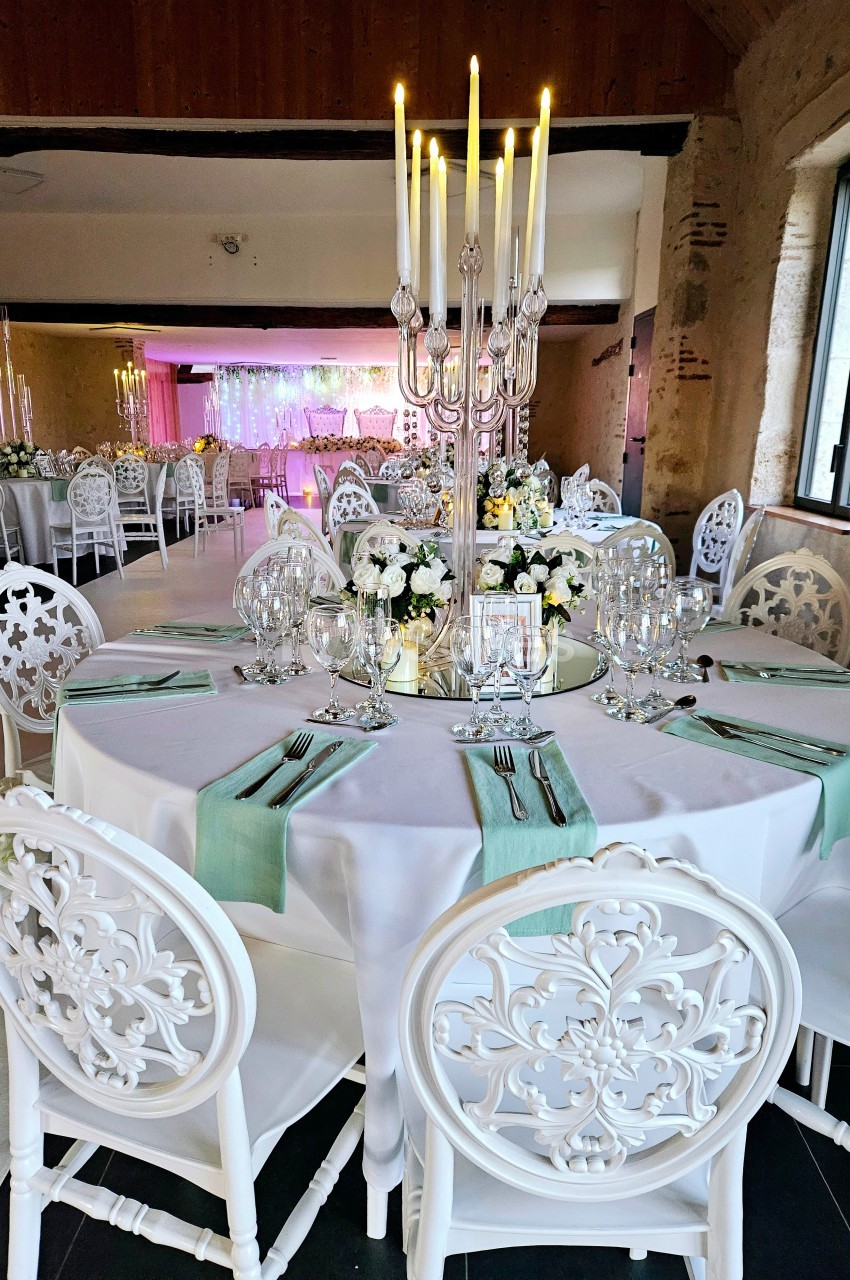 Salle de réception décorée avec des tables rondes, nappes blanches, chaises sculptées et chandeliers élégants.