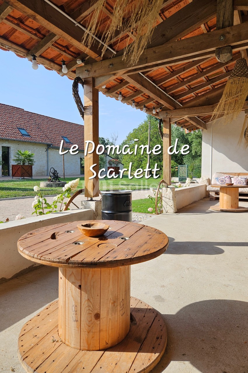Table basse en bois sous une terrasse couverte, avec vue sur un jardin et des bâtiments en arrière-plan.