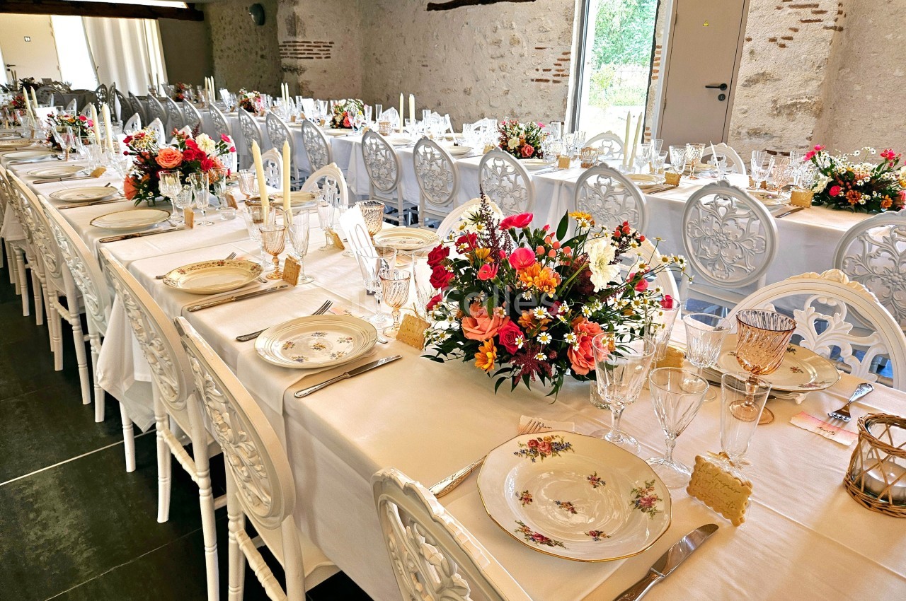 Tables de réception élégamment dressées avec nappes blanches, vaisselle fleurie et bouquets colorés dans une salle en pierre.
