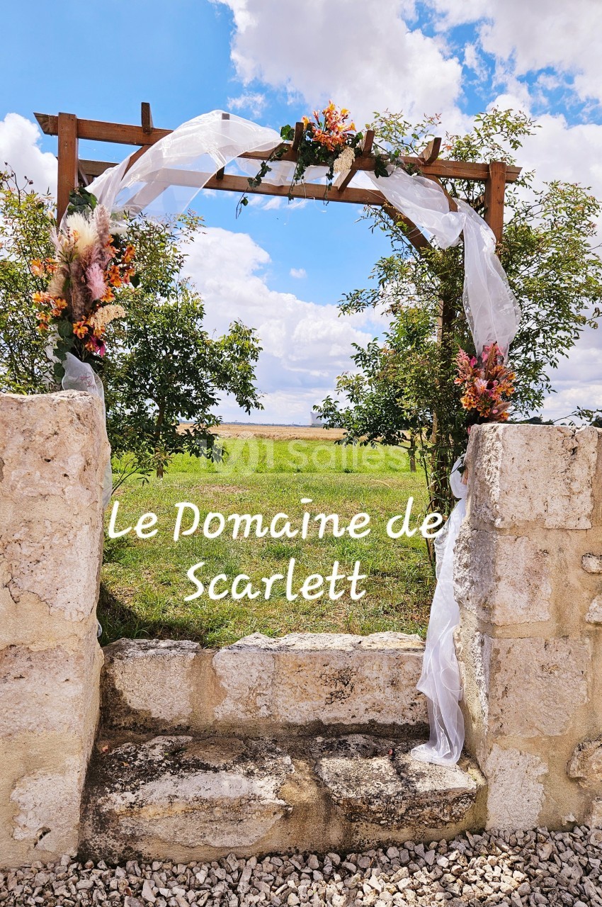 Arche en bois décorée de fleurs et de voilages blancs, encadrée par des murs en pierre, avec un champ en arrière-plan.