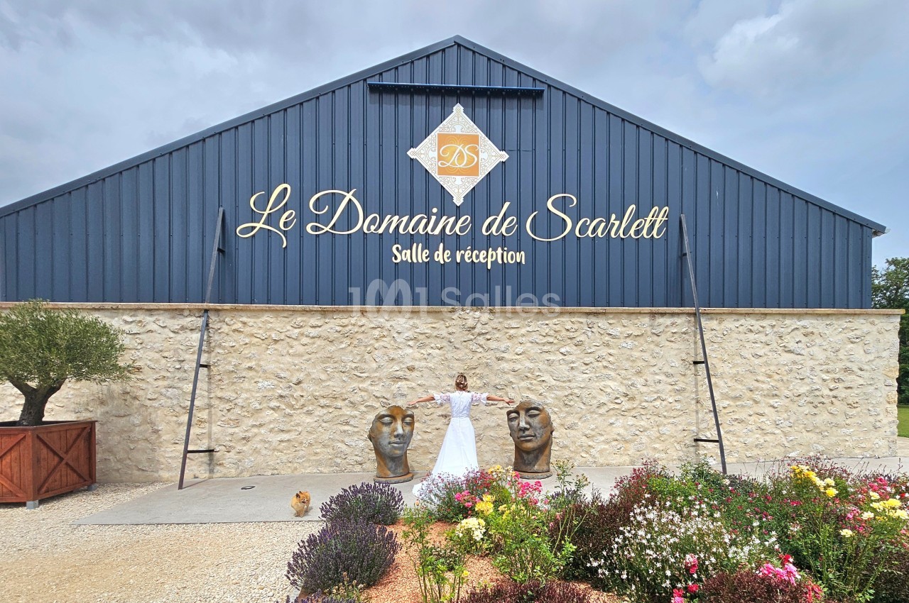 Façade d'une salle de réception en pierre et métal bleu, avec une femme en robe blanche devant des sculptures et des fleurs.