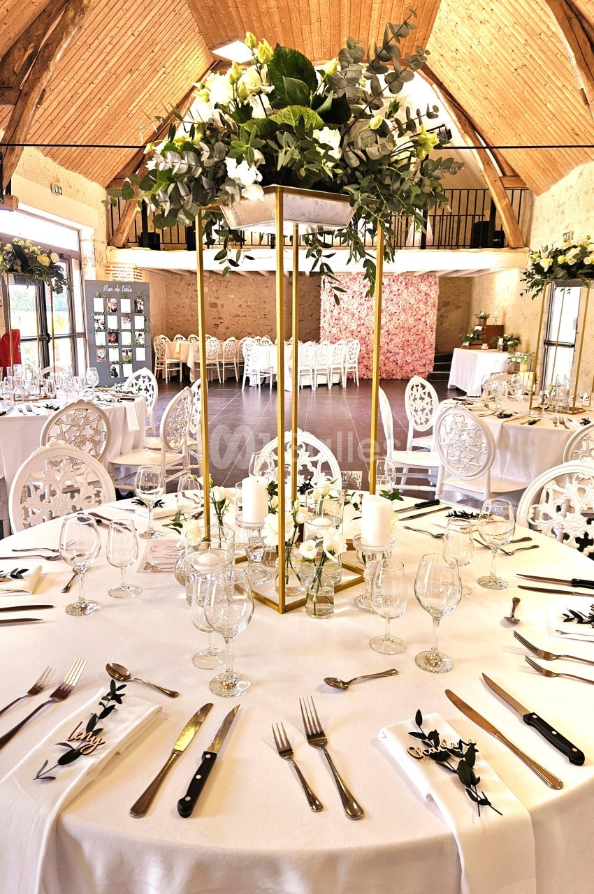 Salle de réception décorée pour un mariage avec tables rondes dressées, centres de table fleuris et chaises blanches.