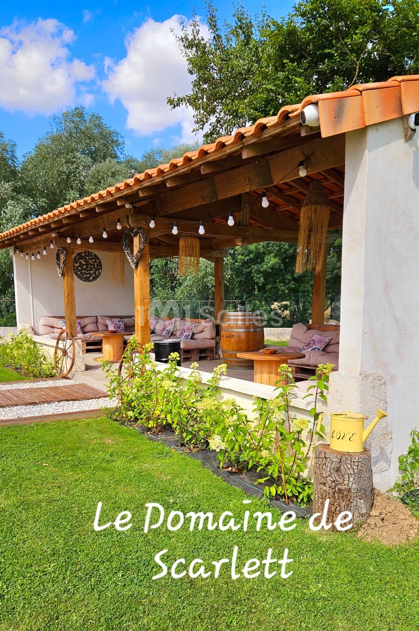 Terrasse couverte avec canapés, décor rustique et végétation, dans un jardin sous un ciel bleu.