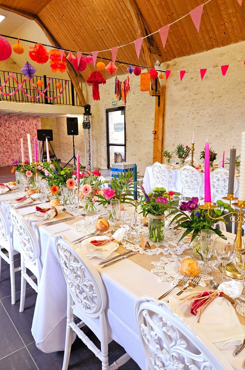 Grande table décorée pour un événement, ornée de fleurs colorées, bougies et guirlandes dans une salle lumineuse.
