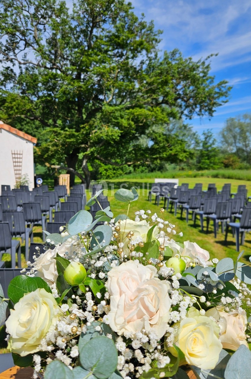 Bouquet de fleurs blanches et vertes au premier plan, avec des chaises disposées en extérieur sous un grand arbre.