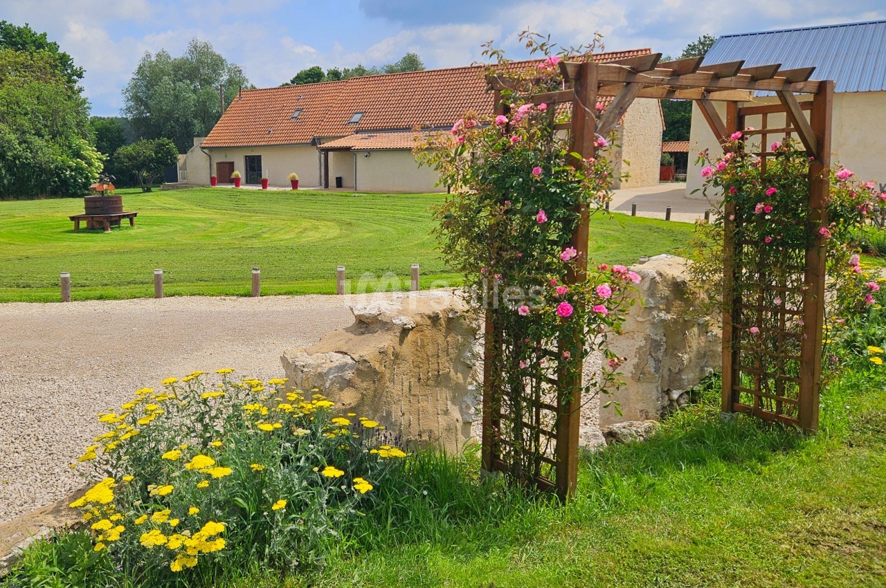 Arche en bois ornée de fleurs devant un jardin verdoyant avec une maison en arrière-plan sous un ciel dégagé.