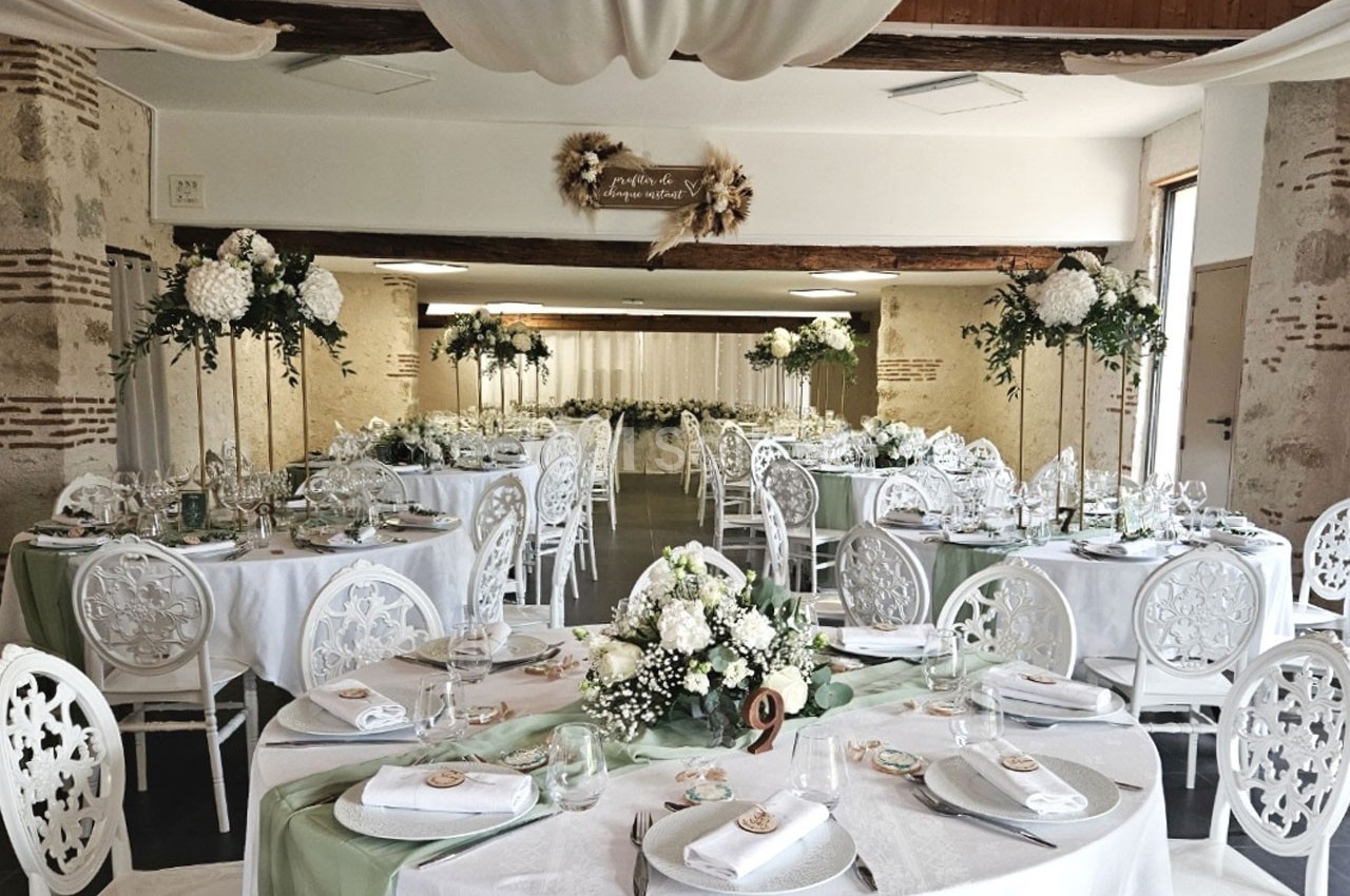 Salle de réception décorée pour un mariage, avec tables rondes, nappes blanches, fleurs blanches et touches de vert.