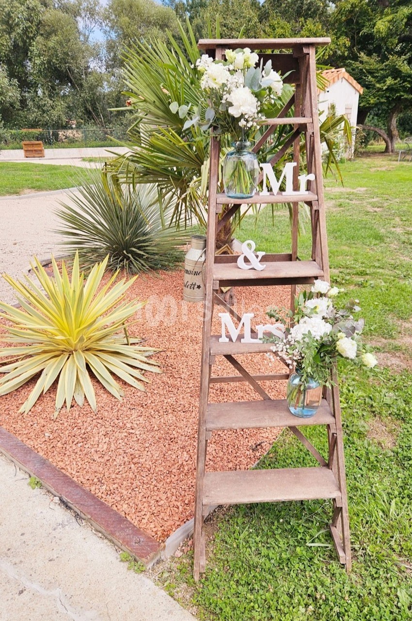 Échelle en bois décorée de fleurs blanches et de lettres ’Mr & Mrs’ dans un jardin avec plantes et graviers.