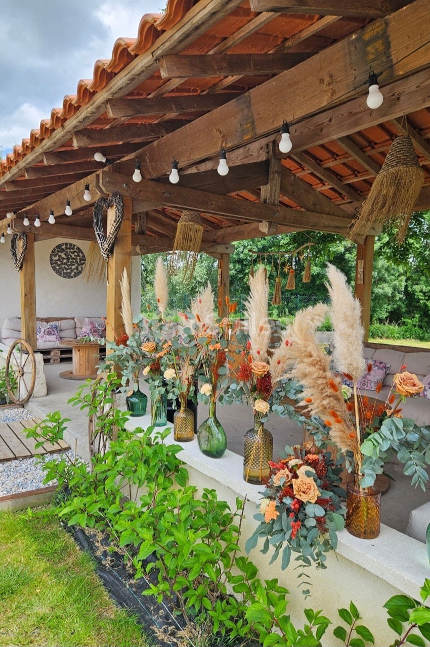 Terrasse couverte en bois décorée de vases fleuris, pampas et guirlandes lumineuses, entourée de verdure.