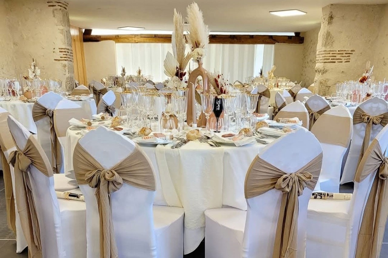Salle de réception décorée avec des tables rondes, nappes blanches, chaises ornées de nœuds beige et centre de table floral.