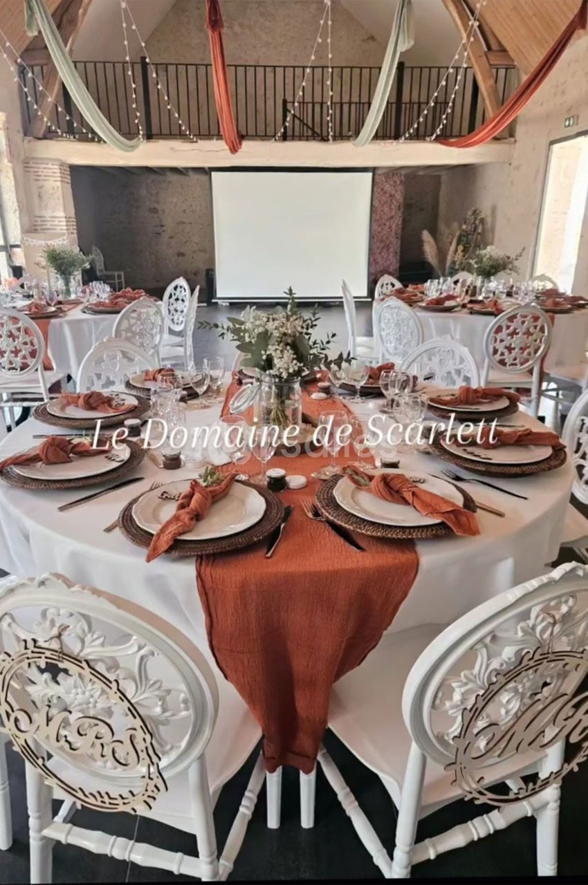 Salle de réception décorée avec des tables rondes, nappes blanches, chemins de table orange et chaises blanches sculptées.